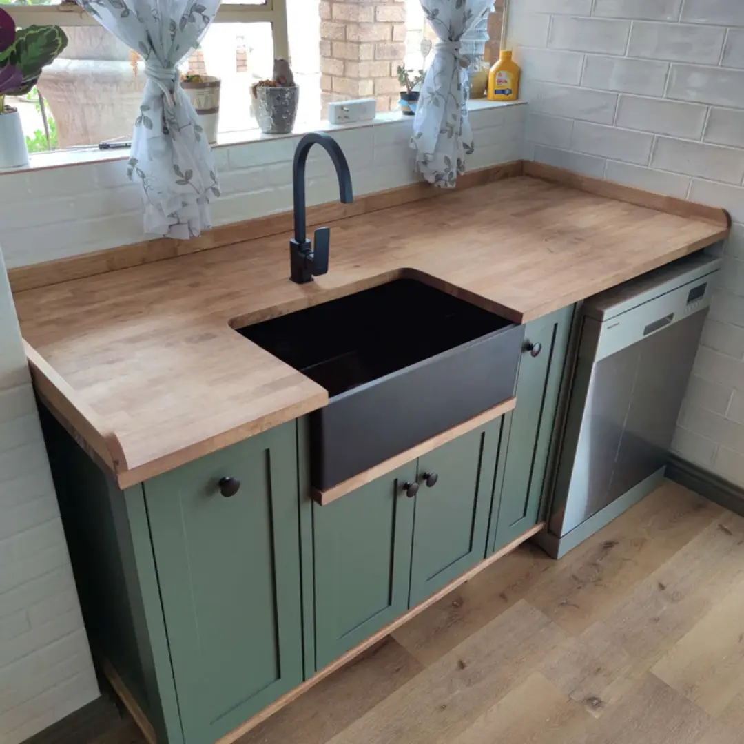 Kitchen sink area with green cabinets, a black farmhouse sink, wooden countertops, stainless steel dishwasher, and sheer floral curtains over a window with natural light.