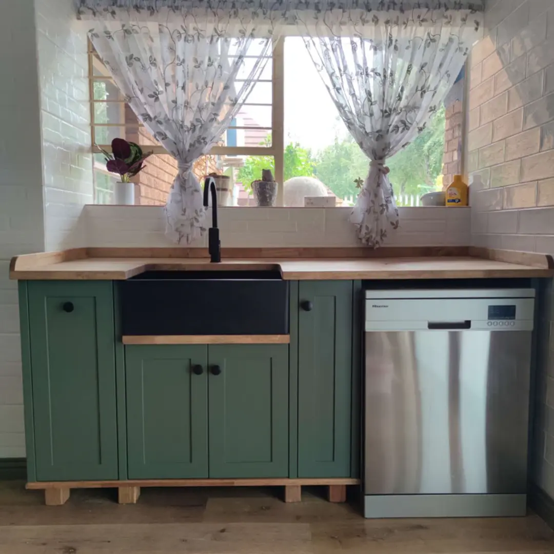 Kitchen sink area with green cabinets, a black farmhouse sink, wooden countertops, stainless steel dishwasher, and sheer floral curtains over a window with natural light.