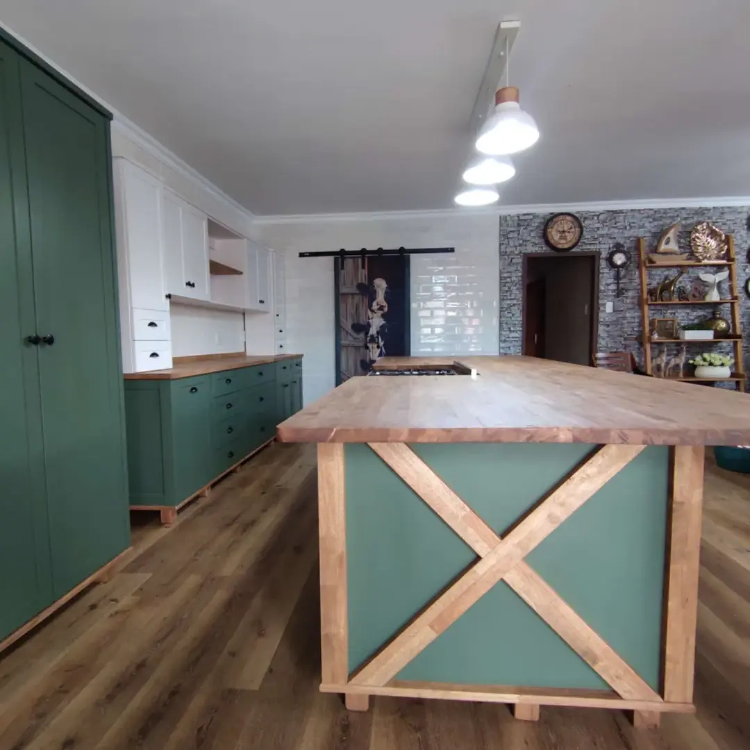 Modern farmhouse kitchen featuring a large wooden island with green paneling and X-frame detail, matching green and white cabinets, wooden countertops, and pendant lighting.