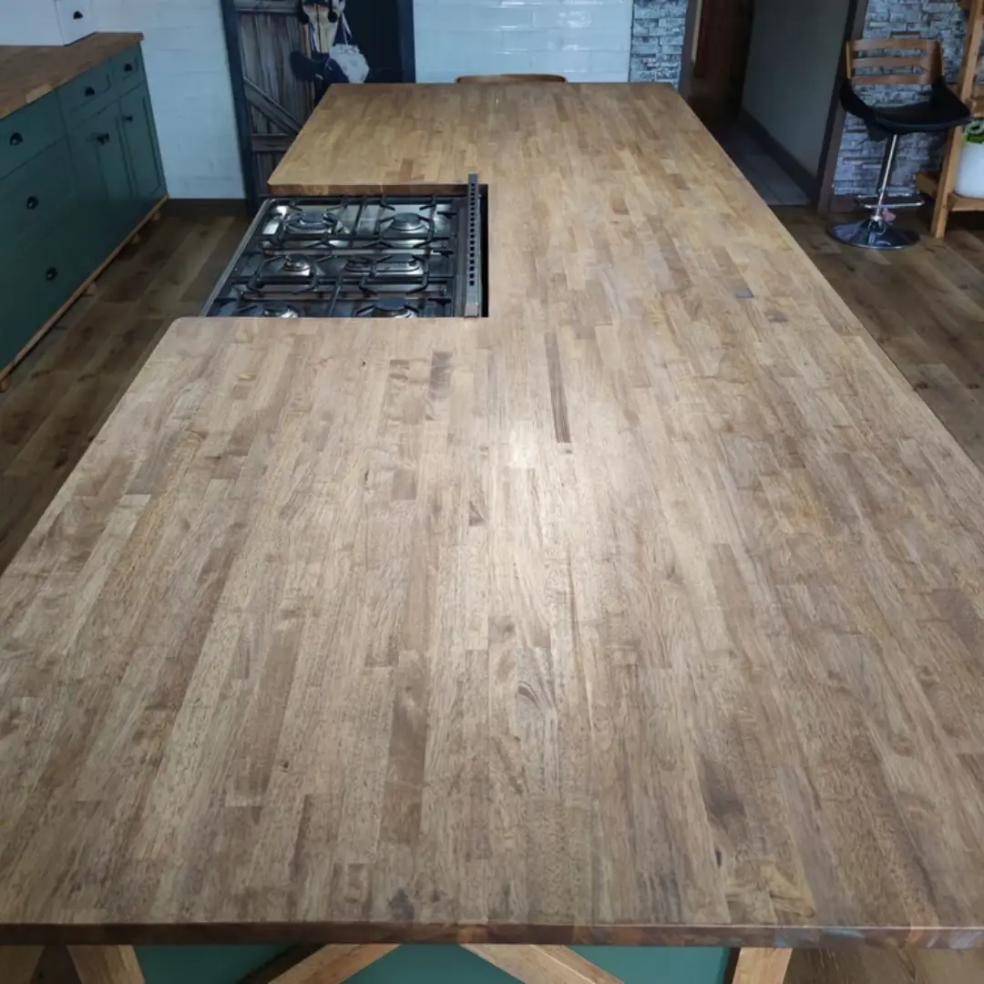 Large wooden kitchen island countertop with built-in gas stove hob, surrounded by green lower cabinets and wooden flooring.