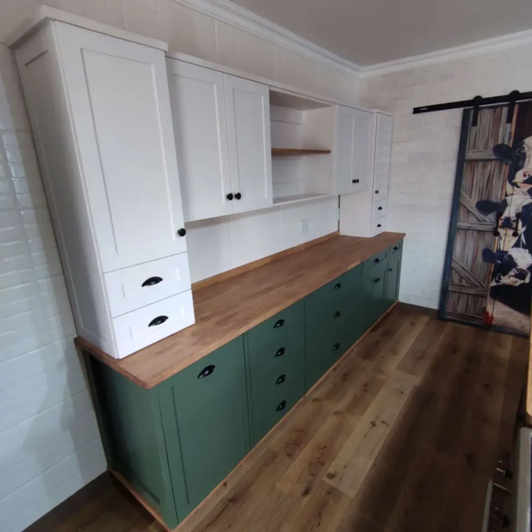 Built-in storage unit with white upper cabinets and green lower cabinets, wooden countertop, black handles, and sliding barn-style door with cow print.