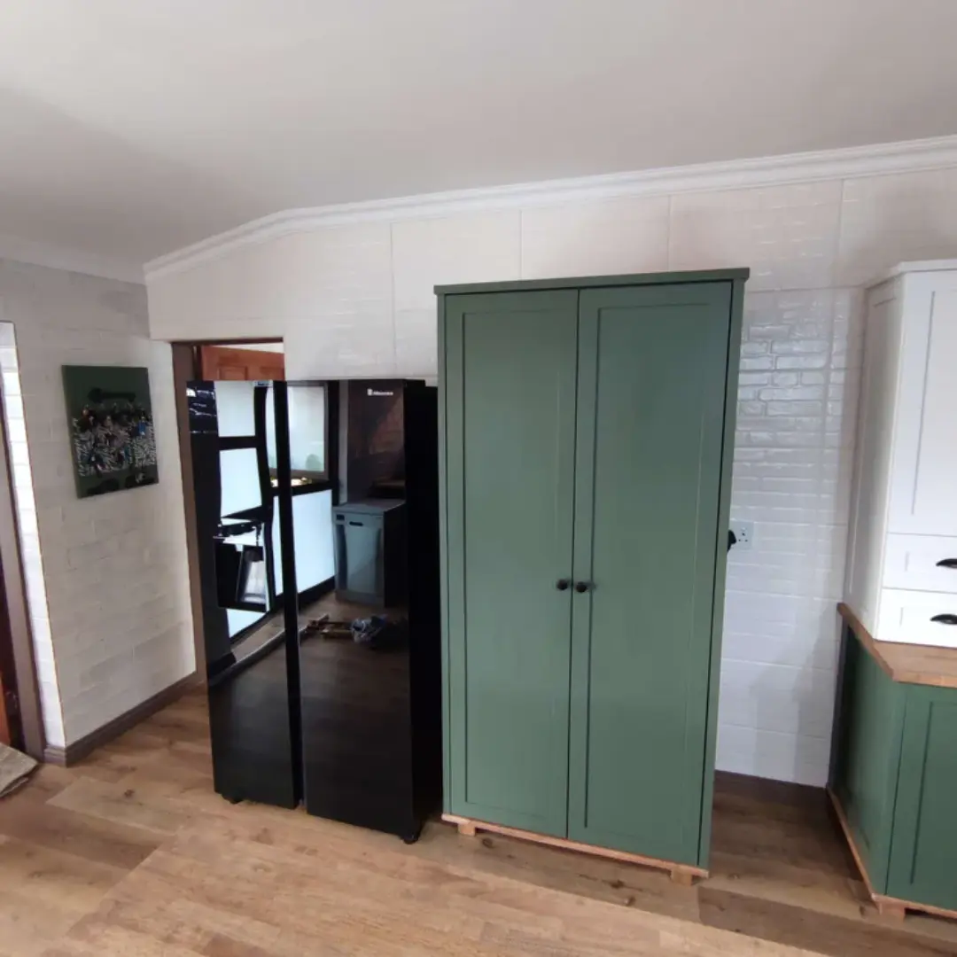 Modern kitchen corner with a black side-by-side fridge next to a tall green pantry cabinet on wooden flooring against a white tiled wall.