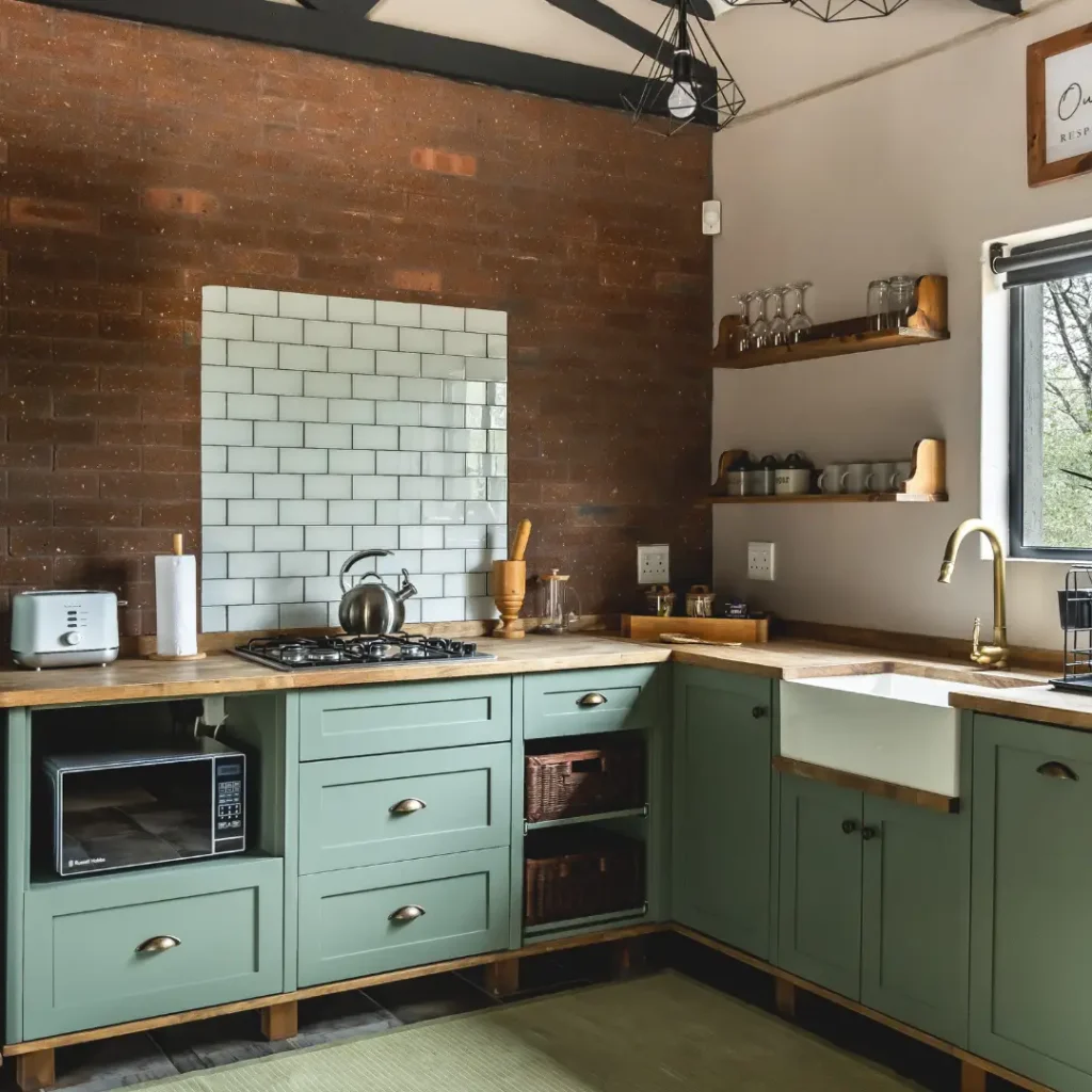 Kitchen with green cabinets, wood countertops, white farmhouse sink, gas stove, and open shelves against a brick wall.