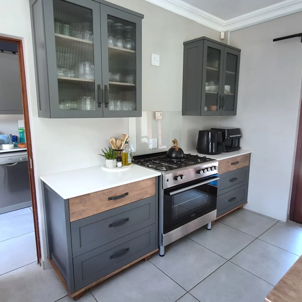 Kitchen with dark grey cabinets, glass-front upper cupboards, white countertop, and stainless steel stove.