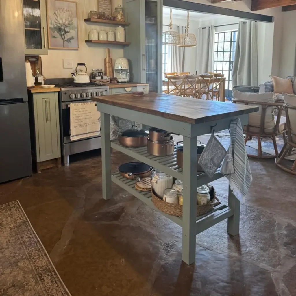 Kitchen with freestanding island table, open shelves storing cookware, green cabinets, and a stove in the background.