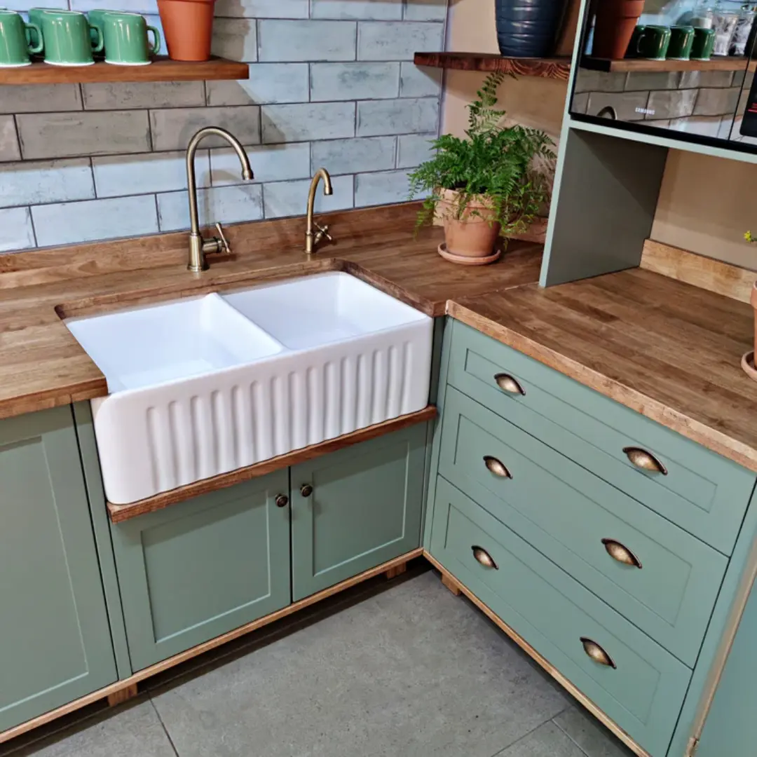 Sage green kitchen cabinets with wooden countertop, farmhouse-style double sink, brass faucets, and rustic open shelving with potted plants and kitchenware.