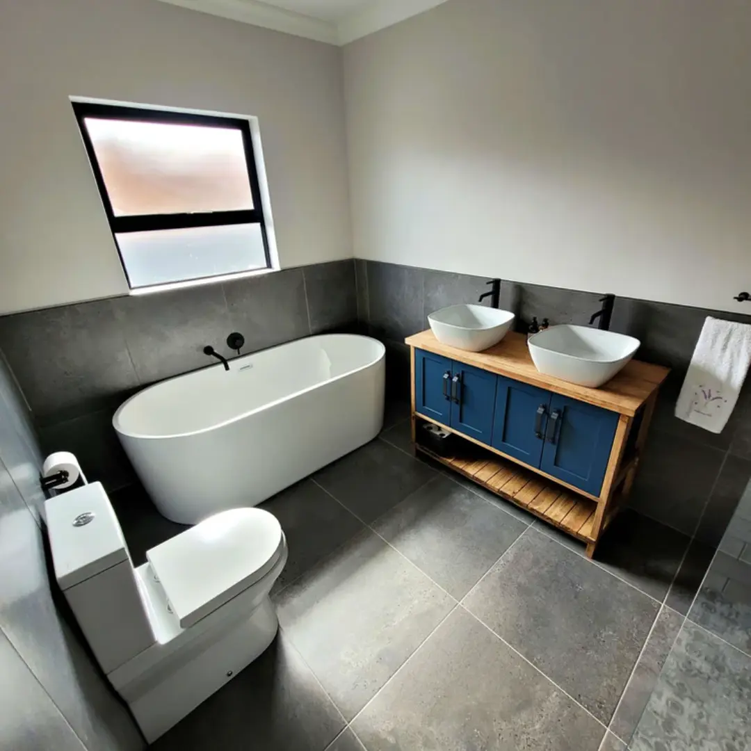 Modern bathroom with a freestanding white bathtub, dark gray floor and wall tiles, and a wooden vanity with blue cabinet doors and two white vessel sinks with black taps.