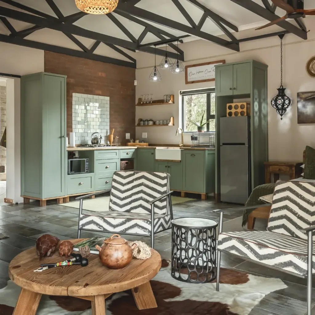 Open-plan kitchen and living area with green cabinets, white farmhouse sink, patterned chairs, and exposed ceiling beams.