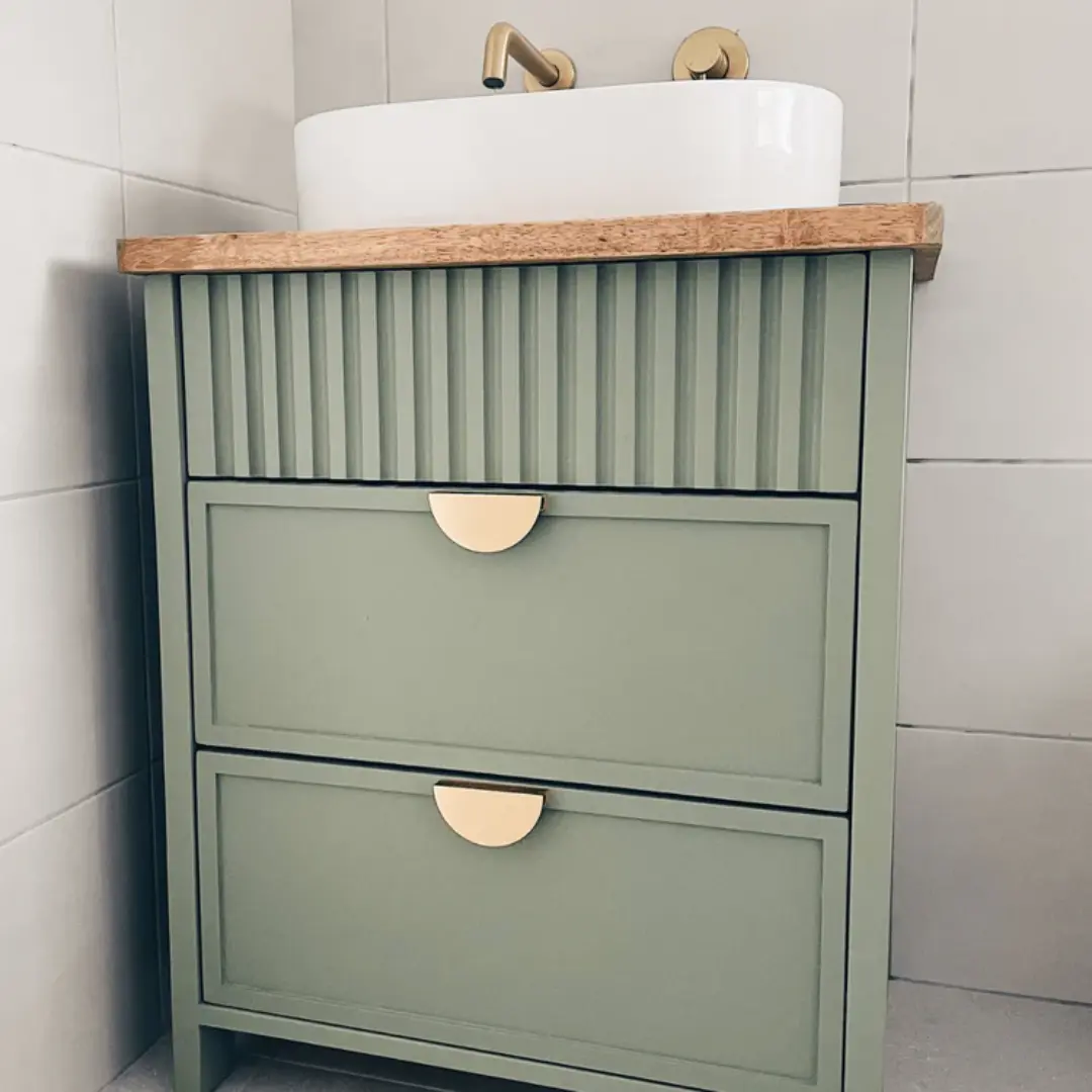 Sage green bathroom vanity with vertical fluted detailing, two shaker-style drawers, brass half-moon handles, wooden countertop, and a white round basin.