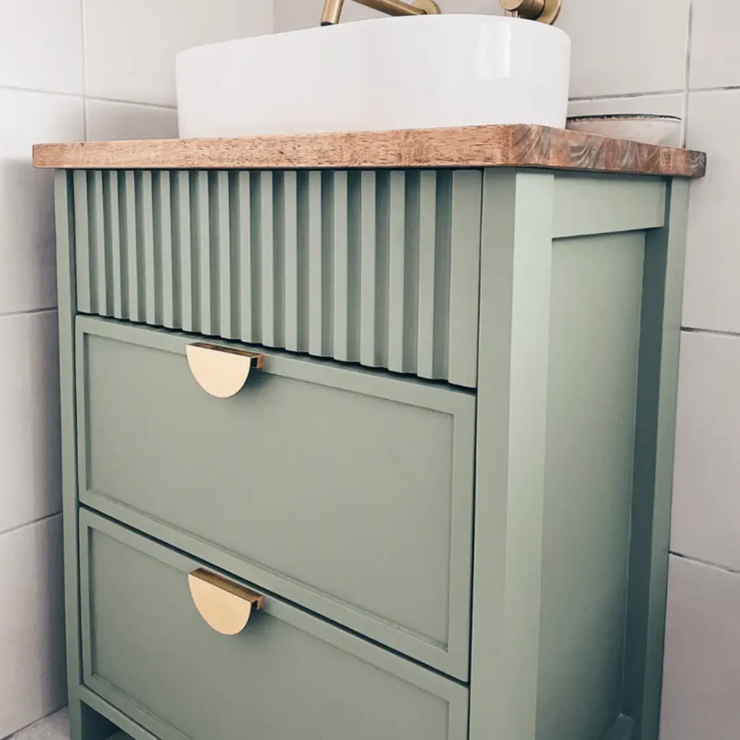 Sage green bathroom vanity with vertical fluted detailing, two shaker-style drawers, brass half-moon handles, wooden countertop, and a white round basin.