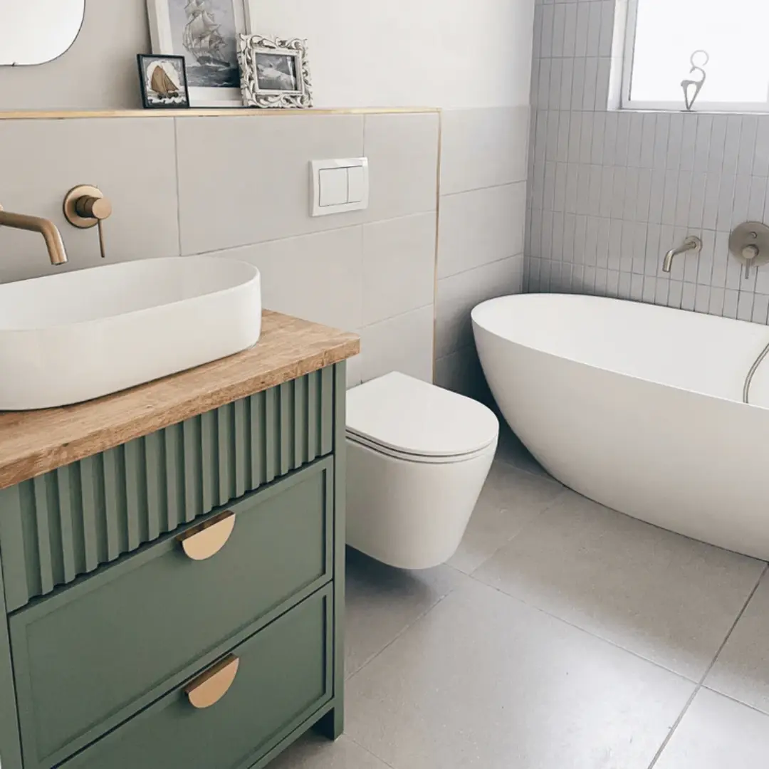 Modern bathroom with a sage green fluted vanity, round white countertop basin, wall-mounted brass tap, wall-hung toilet, and a white freestanding bathtub against light grey tiles.