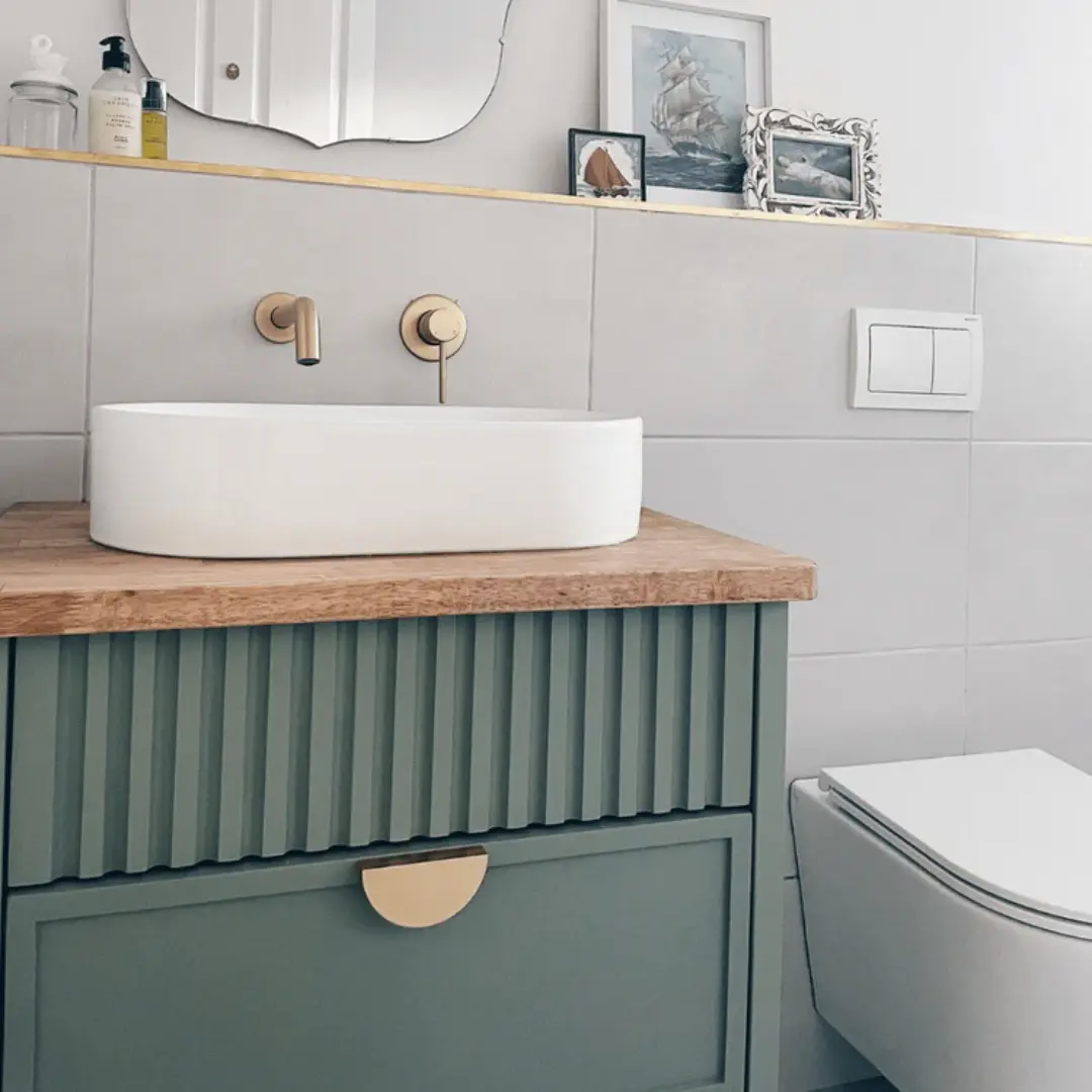Sage green bathroom vanity with vertical fluted detailing, two shaker-style drawers, brass half-moon handles, wooden countertop, and a white round basin.