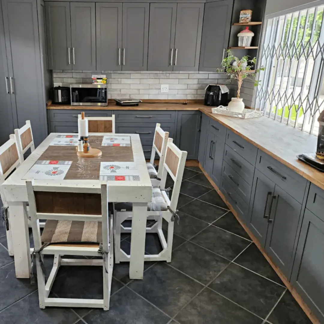 A modern kitchen with matte charcoal grey cabinets, wooden countertops, subway tile backsplash, and a white rustic dining table with matching wooden chairs on a dark tiled floor.