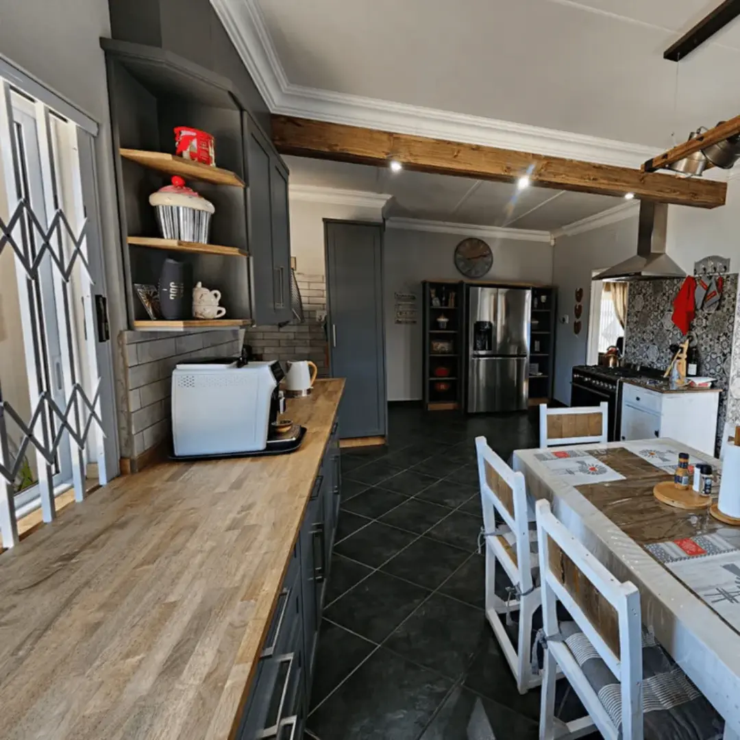 Kitchen interior featuring charcoal grey cabinets, wooden countertops, a patterned backsplash behind the stove, and a white rustic dining table with wooden chairs. A wooden ceiling beam with spotlights is visible overhead, and a stainless steel fridge stands against the back wall.