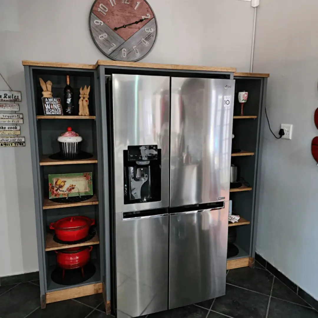Stainless steel double-door fridge built into a grey kitchen cabinet unit with open wooden shelves displaying cookware and décor items, finished with a wood countertop and wall clock above.