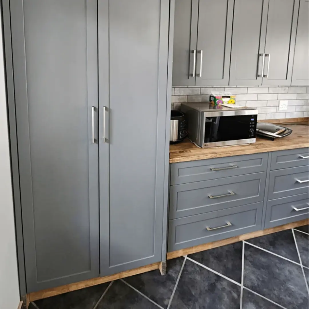 Grey shaker-style kitchen cupboards with stainless steel handles, wood countertop, microwave, and tiled backsplash on dark tiled flooring.