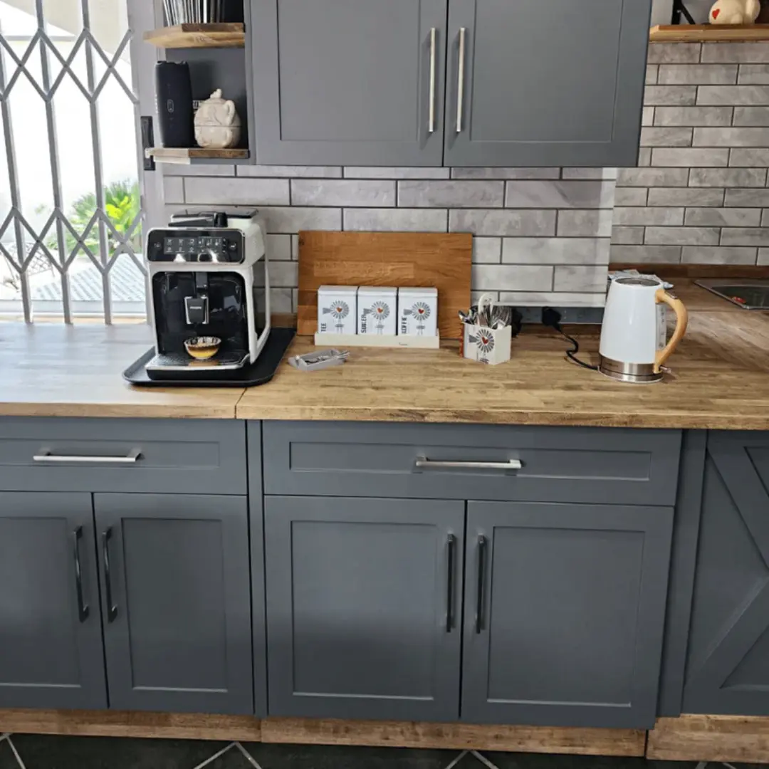 Grey shaker-style kitchen cabinets with stainless steel handles, wood countertop, white coffee machine, white kettle, and light grey subway tile backsplash.