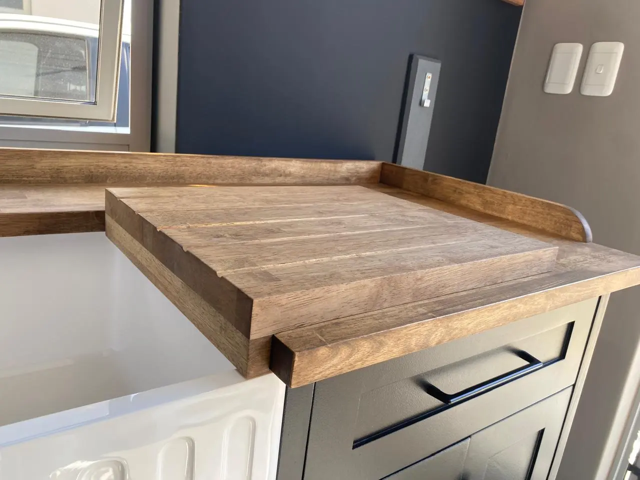 Groovy Panel Butler Sink Showroom Left Kitchen sink area with green cabinets, a black farmhouse sink, wooden countertops, stainless steel dishwasher, and sheer floral curtains over a window with natural light.
